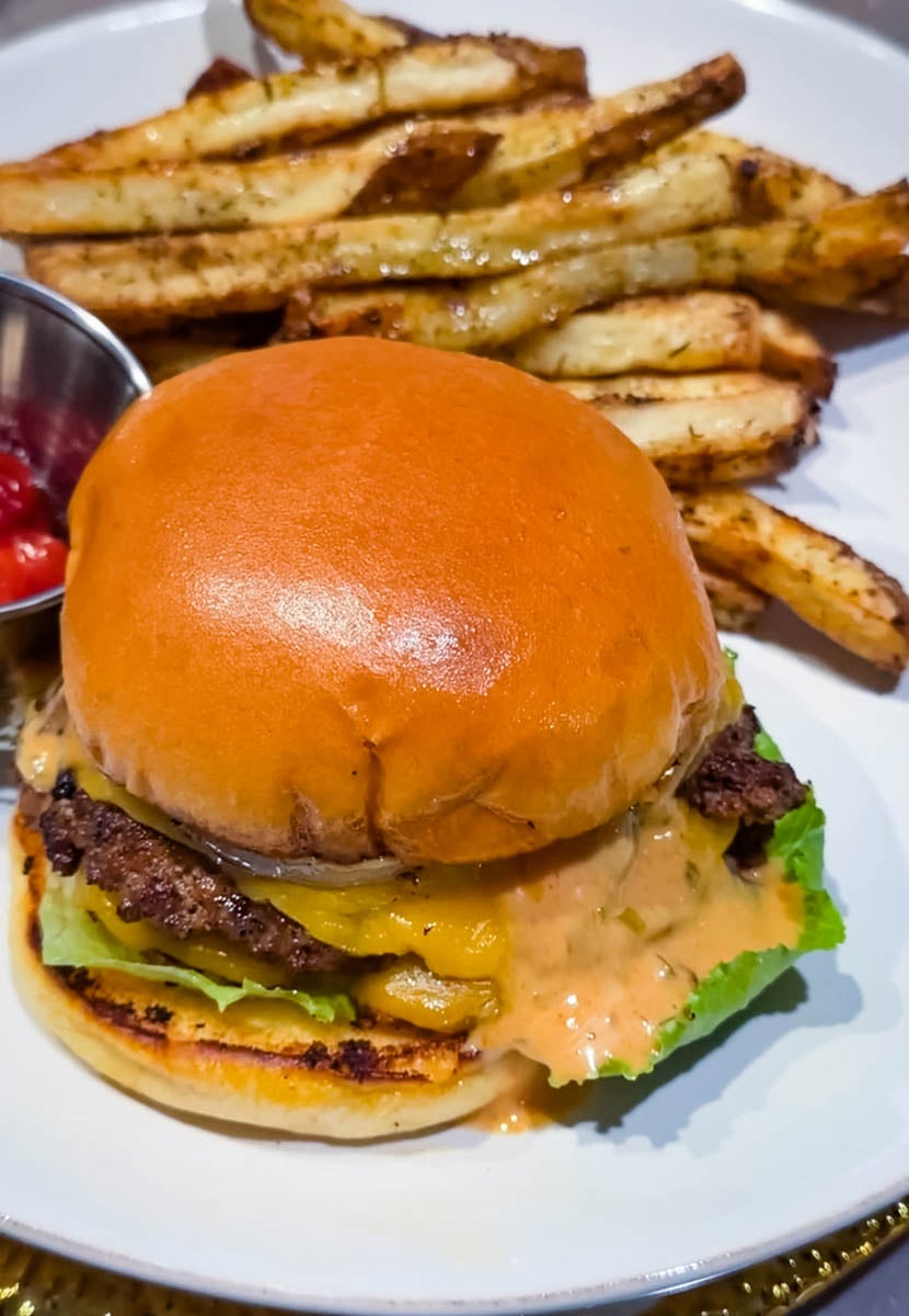 Smash burger topped with melted cheese, lettuce, pickles, onions, and burger sauce on a toasted brioche bun, served with seasoned fries and ketchup.