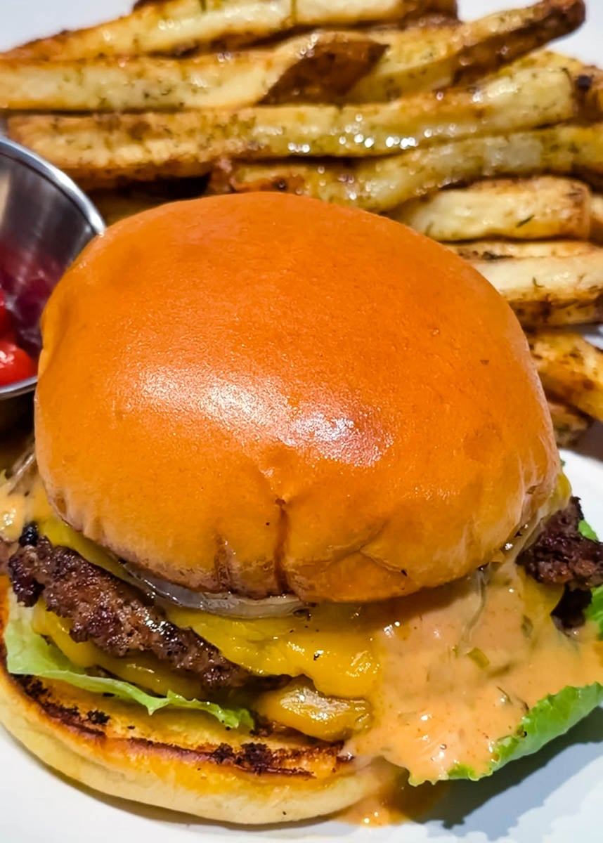 Close-up of smash burger with melted cheese, lettuce, pickles, onions, and creamy burger sauce on a toasted brioche bun with fries in the background.
