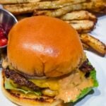 Close-up of a homemade smash burger with crispy beef patties, melted cheese, lettuce, pickles, onions, and burger sauce on a toasted brioche bun.