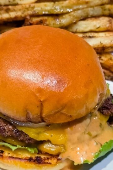 Close-up of a homemade smash burger with crispy beef patties, melted cheese, lettuce, pickles, onions, and burger sauce on a toasted brioche bun.