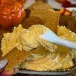 Graham cracker topped with thick pumpkin cheesecake dip and an extra spoonful of dip, with the serving platter and fall decorations in the background.