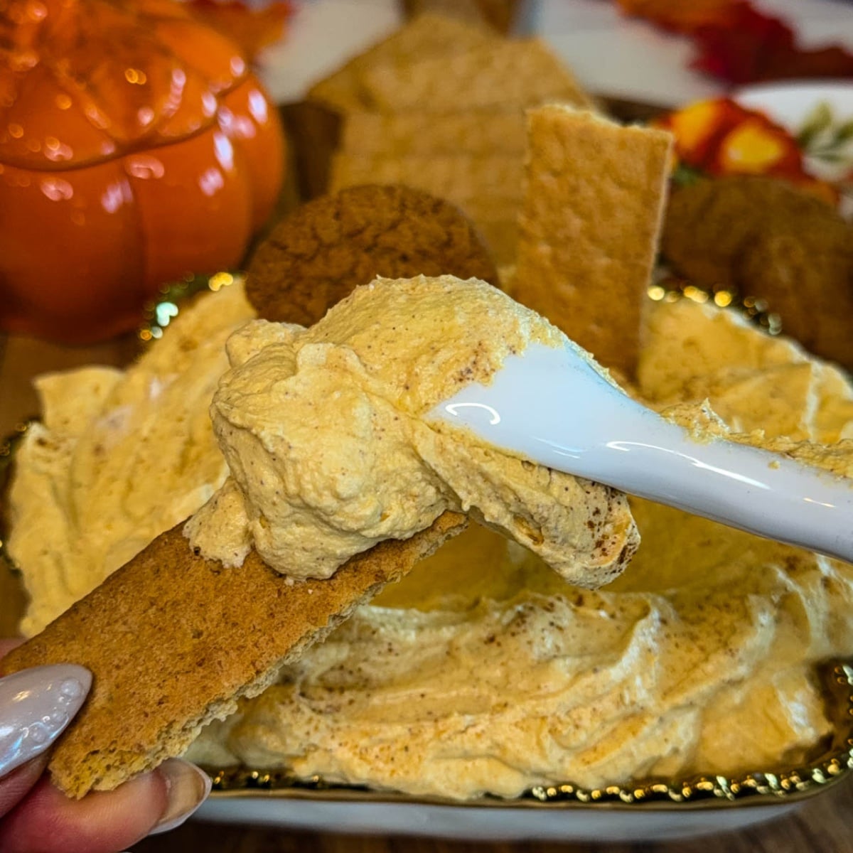 Graham cracker topped with thick pumpkin cheesecake dip and an extra spoonful of dip, with the serving platter and fall decorations in the background.