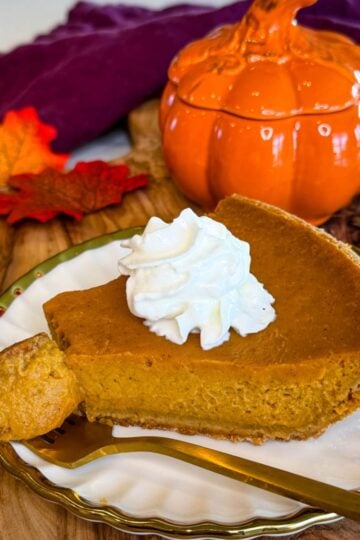 Slice of pumpkin pie with graham cracker crust topped with whipped cream and served with a gold fork.