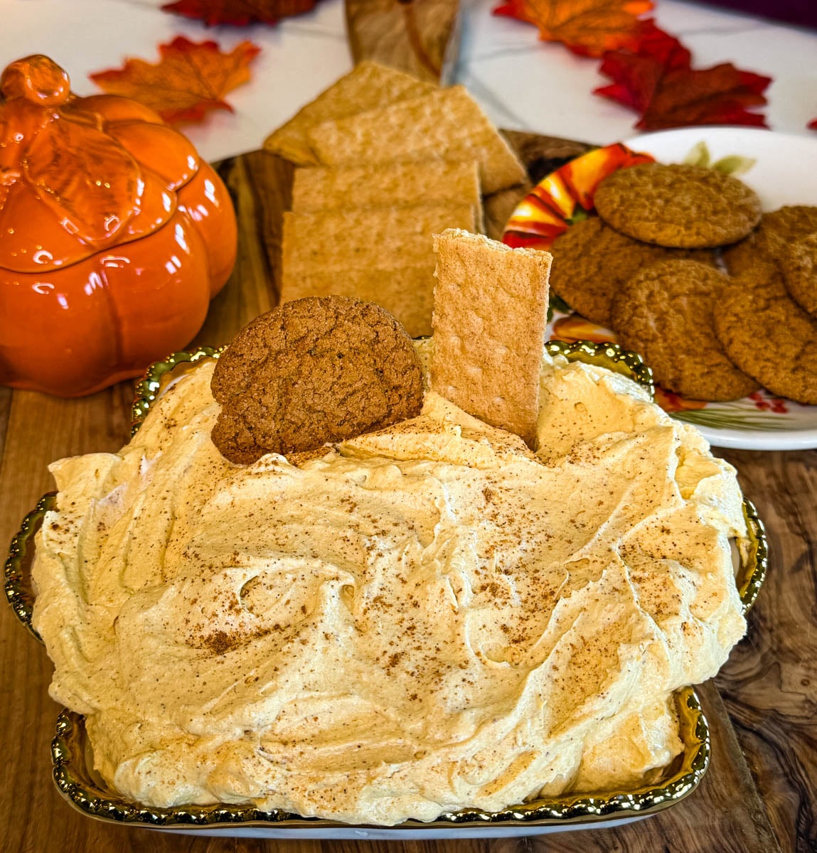 Pumpkin cheesecake dip served on a decorative tray with graham crackers and ginger cookies, surrounded by fall pumpkin and leaf decorations.