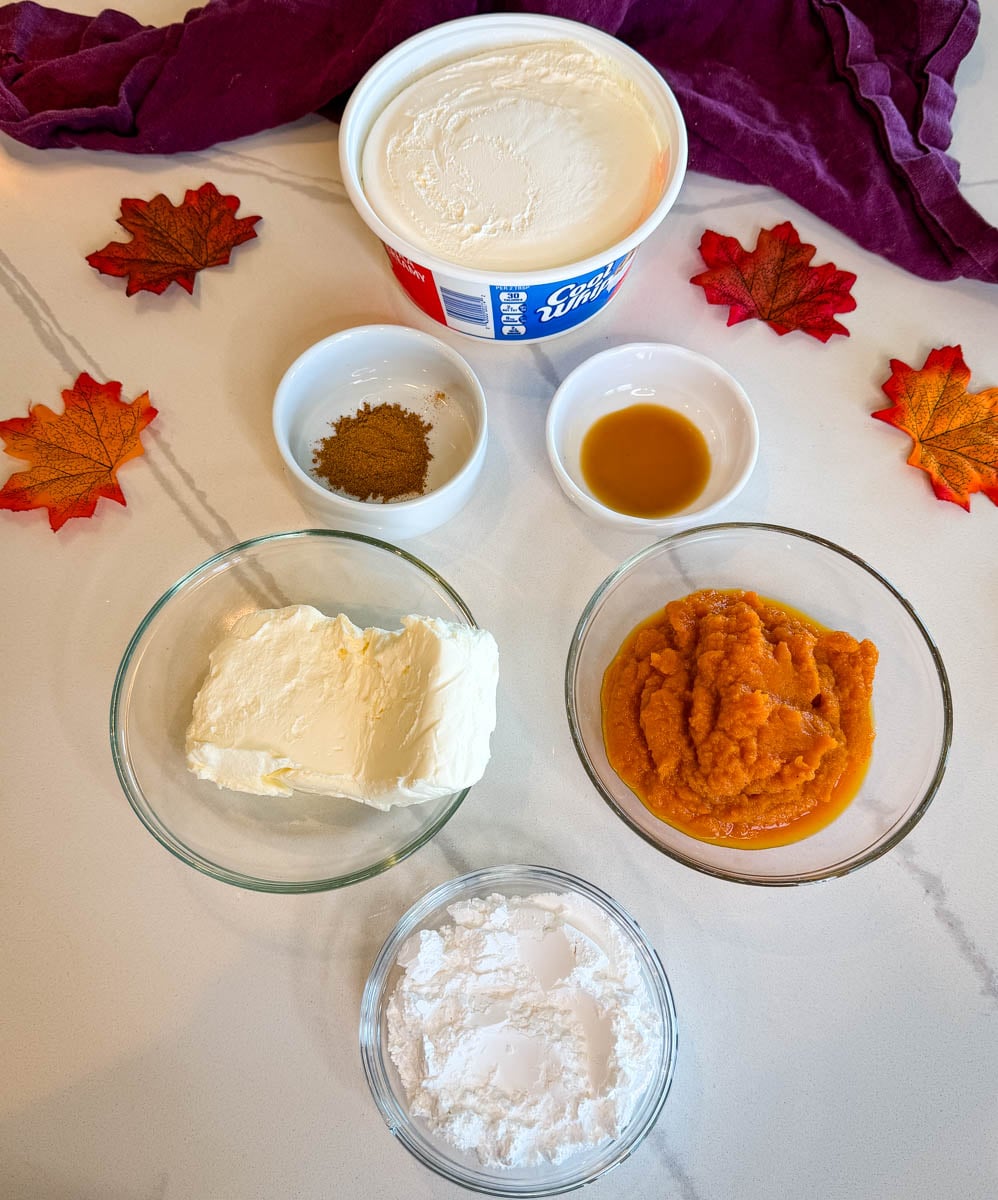 Ingredients for pumpkin cheesecake dip arranged in bowls, including pumpkin puree, cream cheese, powdered sugar, whipped topping, vanilla, and pumpkin pie spice.