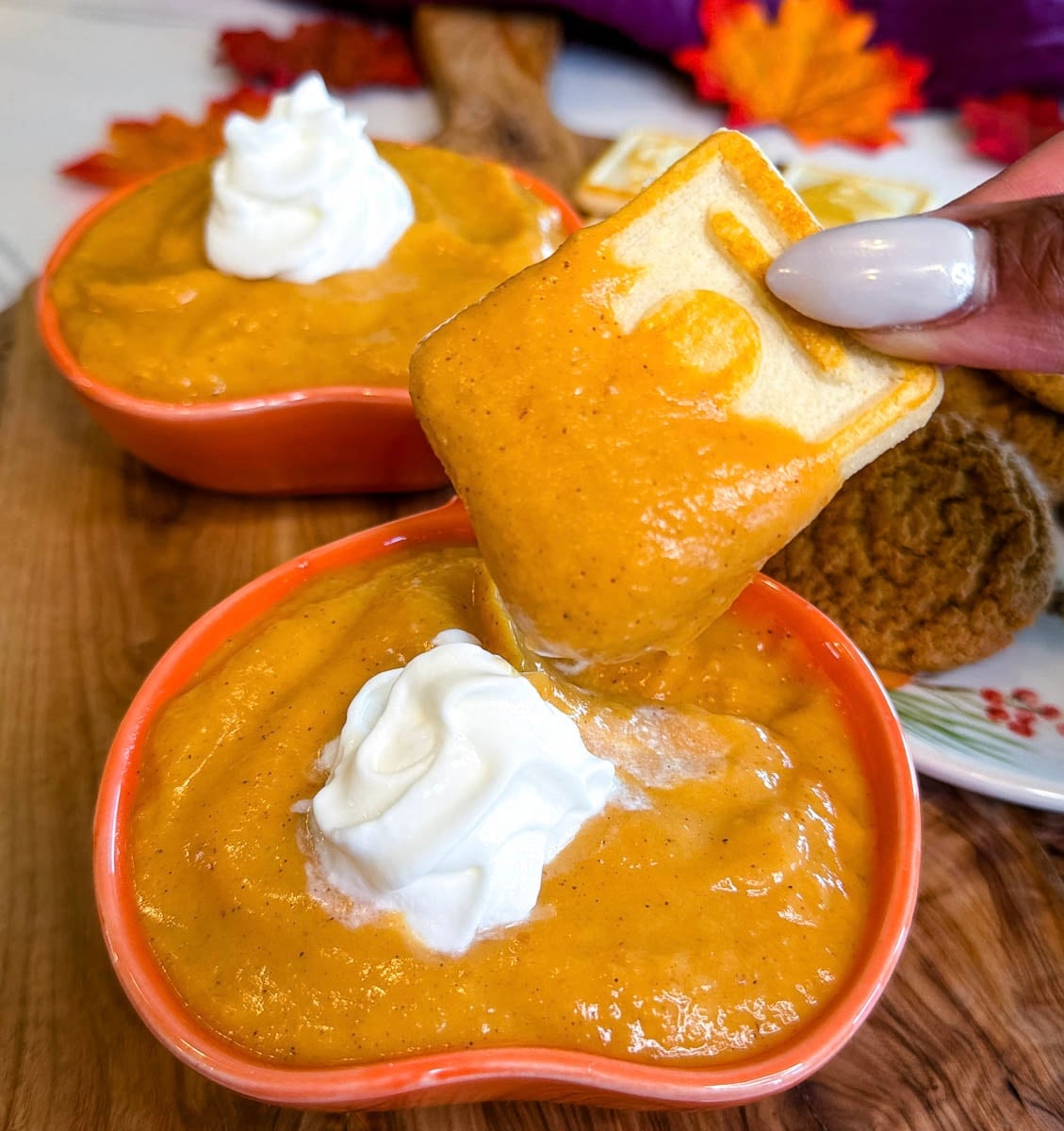 Fall-themed cookie dipped into creamy pumpkin pie dip served in an orange pumpkin-shaped bowl and topped with whipped cream.