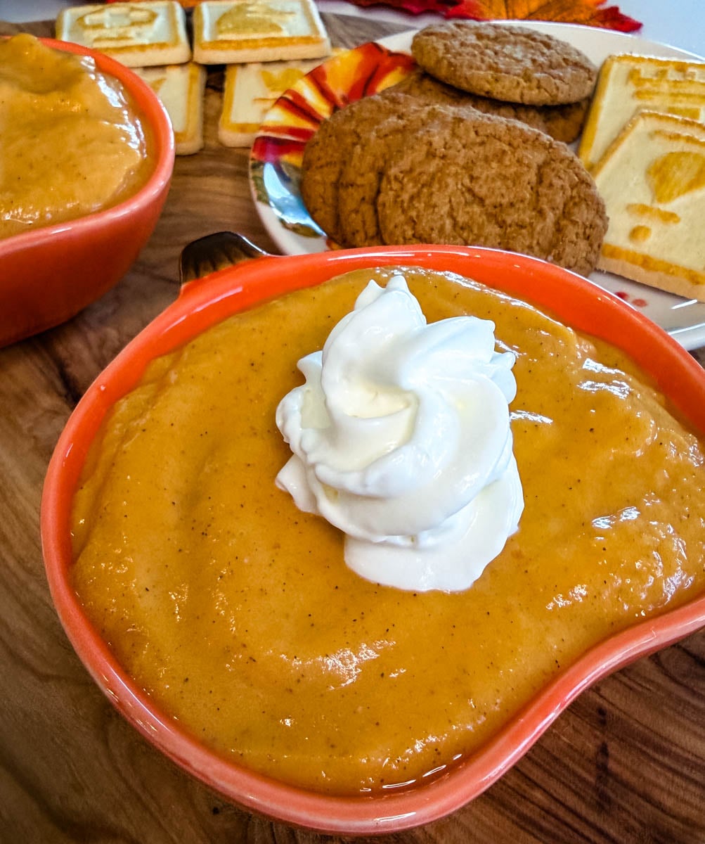Close-up of creamy pumpkin pie dip topped with whipped cream in an orange pumpkin-shaped bowl with cookies in the background.