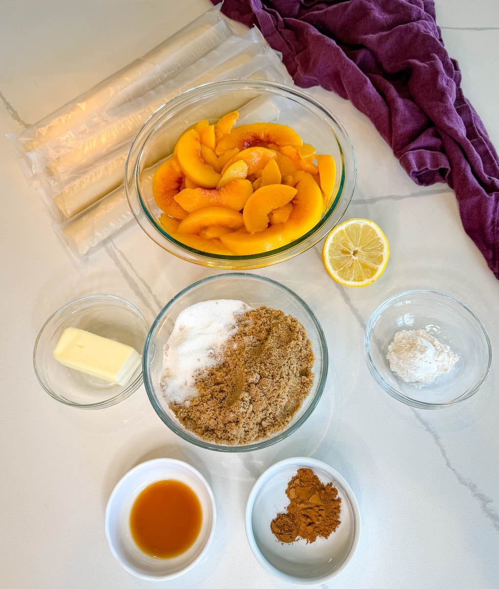 Southern peach cobbler ingredients arranged in bowls, including sliced peaches, pie crust, butter, granulated and brown sugar, flour, lemon, vanilla extract, cinnamon, and nutmeg.