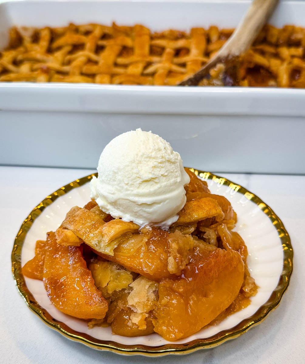 Southern peach cobbler served on a plate with tender cinnamon-spiced peaches, flaky pie crust, and a scoop of vanilla ice cream, with the baked cobbler in the background.