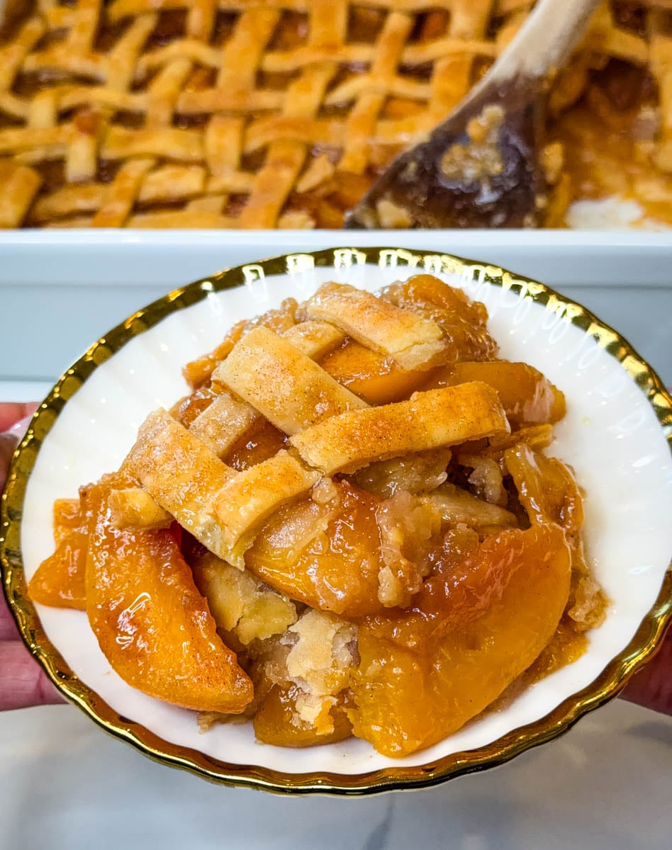 Close-up of Southern peach cobbler with juicy peaches and pieces of golden flaky pie crust served on a white and gold plate.