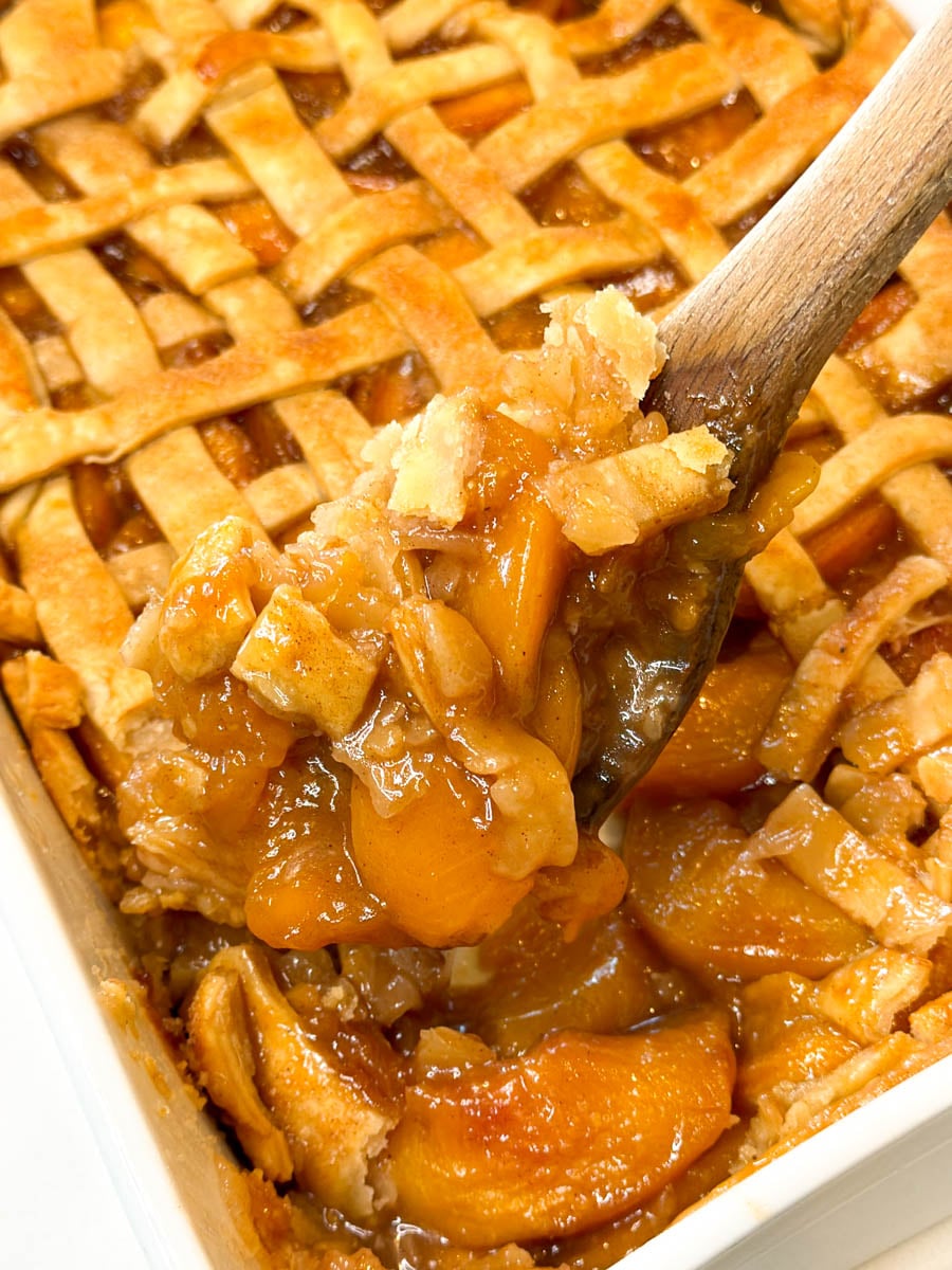 Wooden spoon lifting warm peach cobbler from the baking dish, showing tender peaches, thick cinnamon-spiced filling, and flaky golden pie crust.