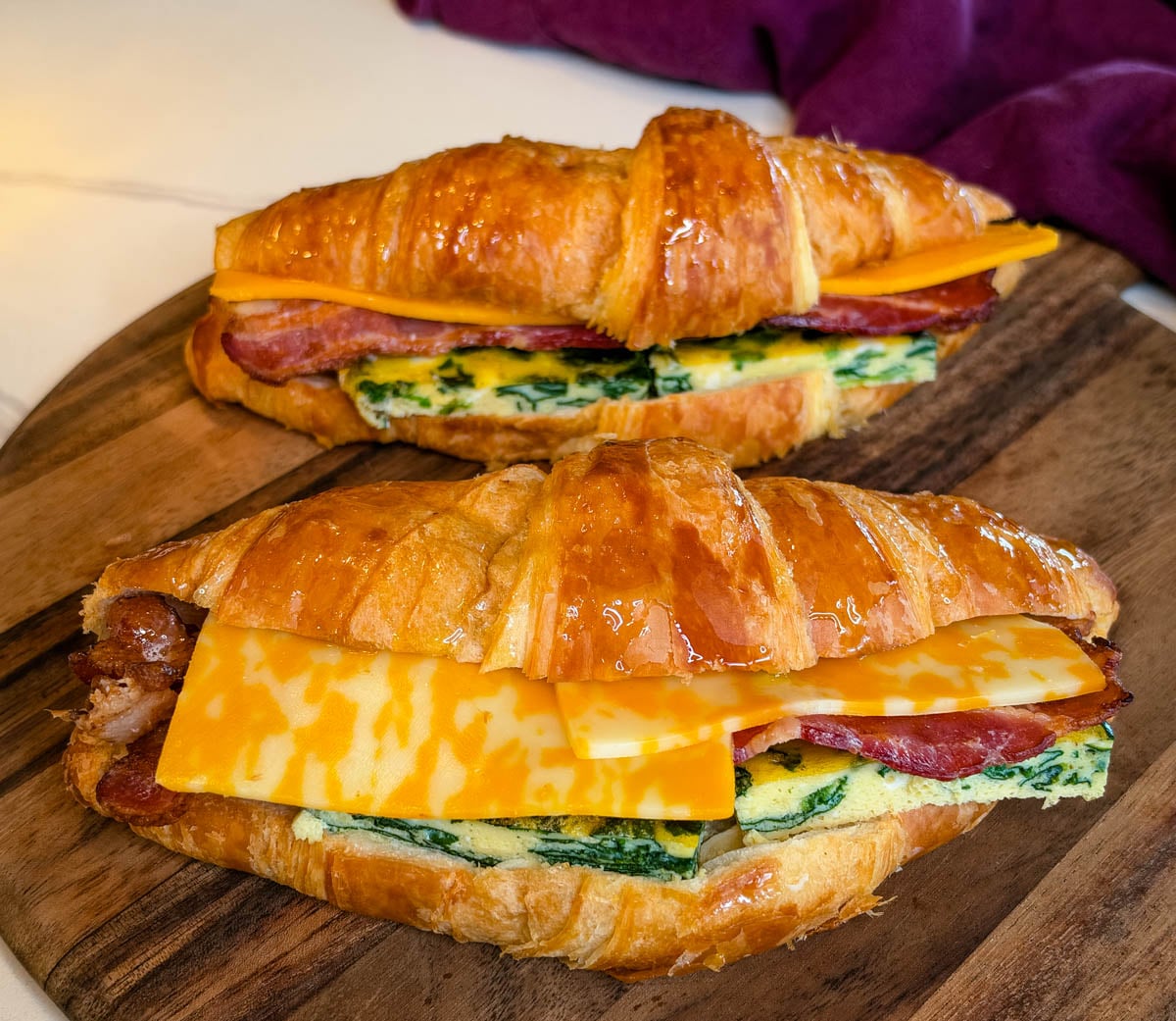 Two croissant breakfast sandwiches filled with spinach eggs, crispy bacon, and sliced cheese, served on a wooden cutting board.