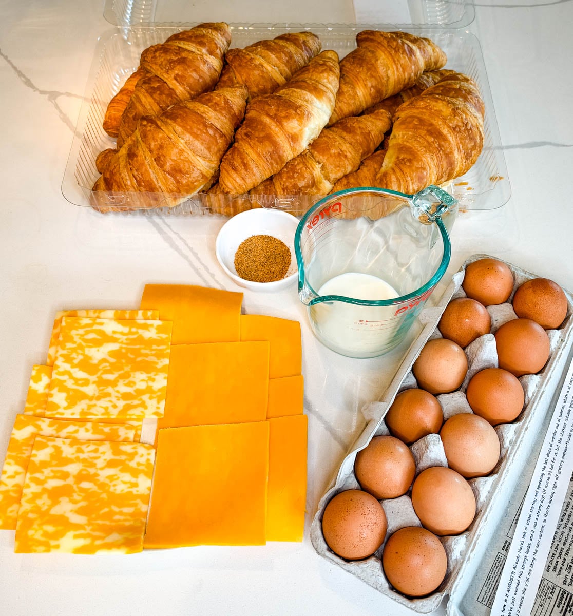 Ingredients for croissant breakfast sandwiches including croissants, eggs, sliced cheddar and Colby Jack cheese, milk, and seasoning arranged on a countertop.