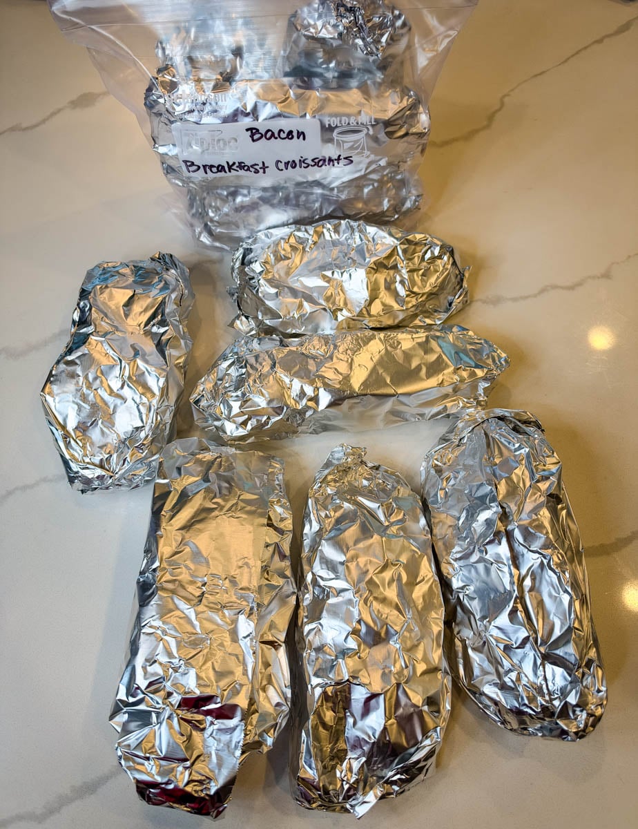 Individually foil-wrapped croissant breakfast sandwiches arranged beside a labeled freezer storage bag for meal prep.