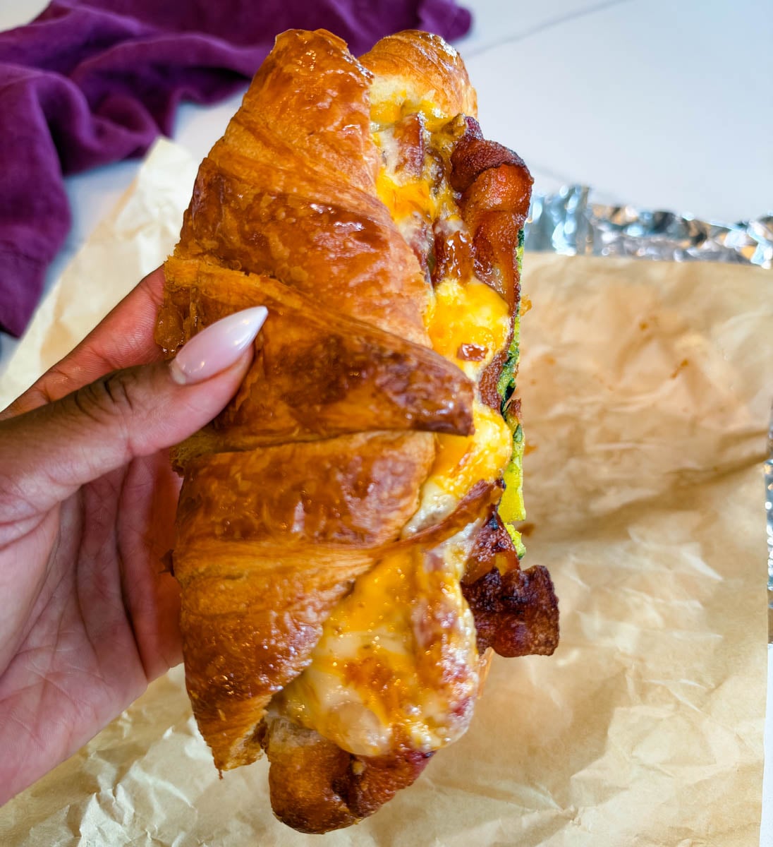 Hand holding a warm croissant breakfast sandwich filled with spinach eggs, crispy bacon, and melted cheese over parchment paper.