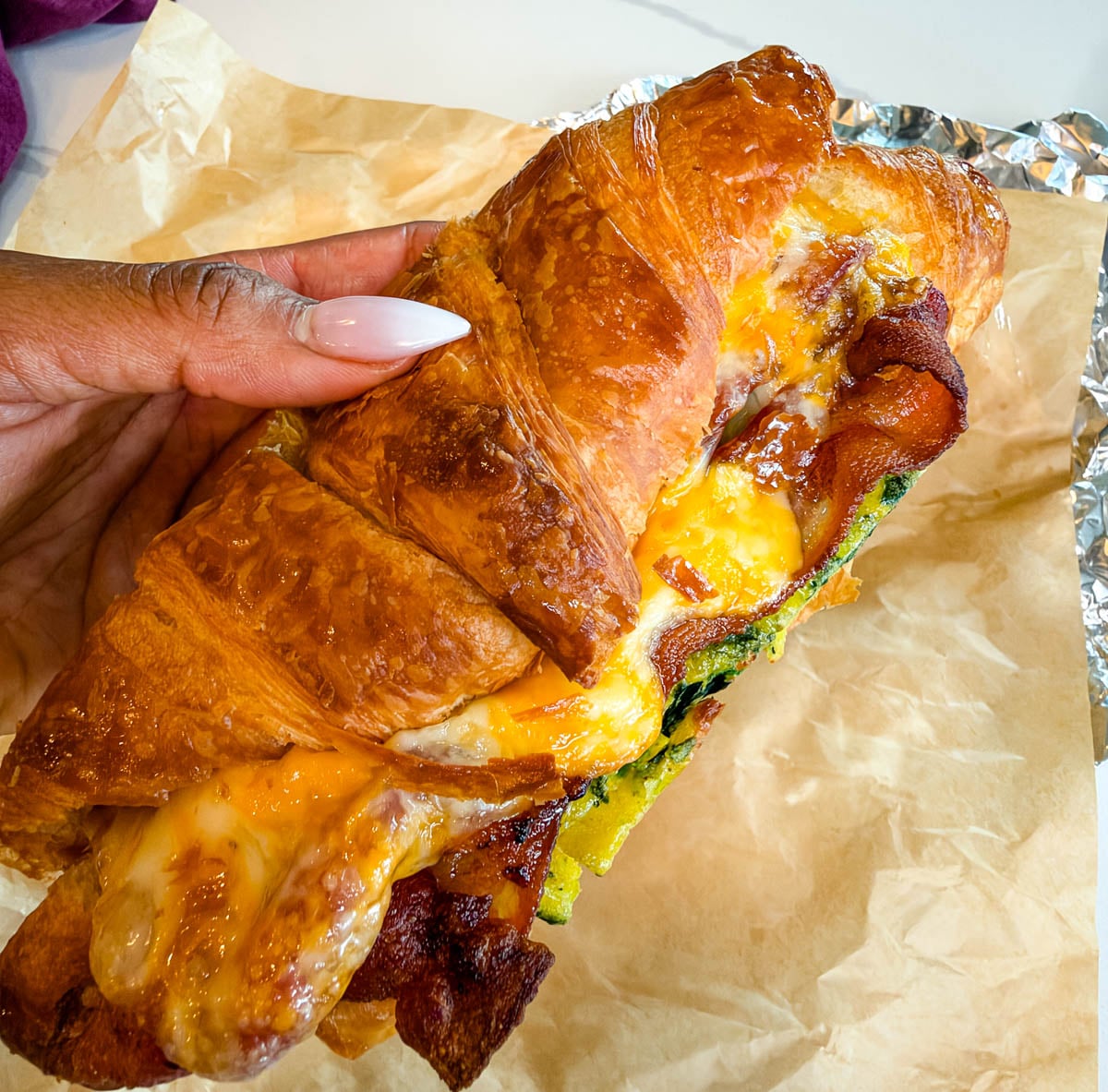Close-up of a croissant breakfast sandwich held in one hand, showing crispy bacon, spinach eggs, and gooey melted cheese between flaky layers.