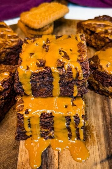 Two thick Biscoff brownie squares stacked on a wooden board, showing fudgy chocolate centers and cookie pieces with melted cookie butter dripping over the brownies.