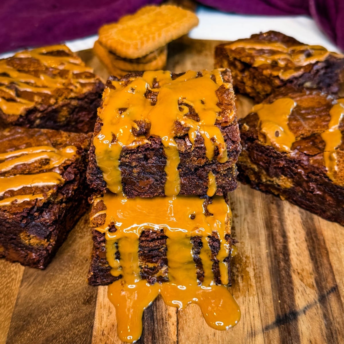 Two thick Biscoff brownie squares stacked on a wooden board, showing fudgy chocolate centers and cookie pieces with melted cookie butter dripping over the brownies.