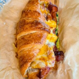 Close-up of a flaky croissant breakfast sandwich filled with crispy bacon, spinach eggs, and gooey melted cheese on parchment paper.
