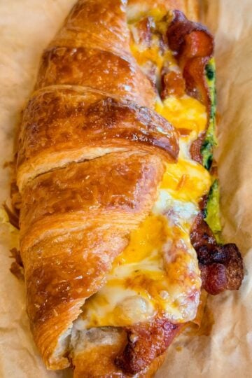 Close-up of a flaky croissant breakfast sandwich filled with crispy bacon, spinach eggs, and gooey melted cheese on parchment paper.
