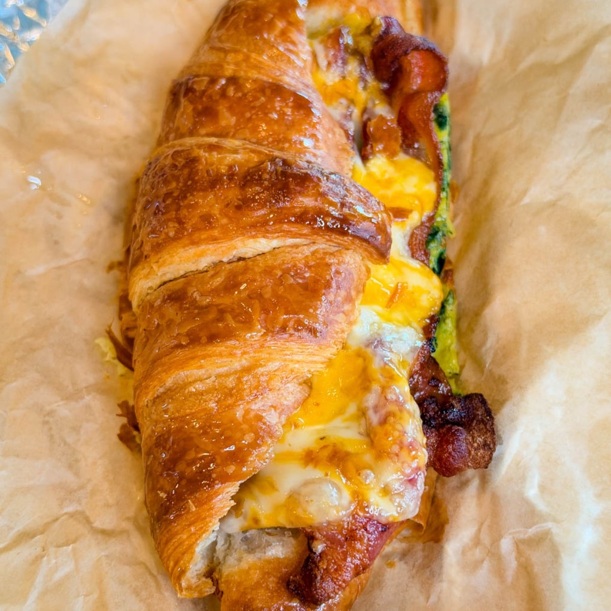 Close-up of a flaky croissant breakfast sandwich filled with crispy bacon, spinach eggs, and gooey melted cheese on parchment paper.