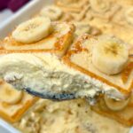Close-up of a spoonful of creamy Chessman banana pudding with layers of banana, pudding, and cookies, with the finished dessert in the background.