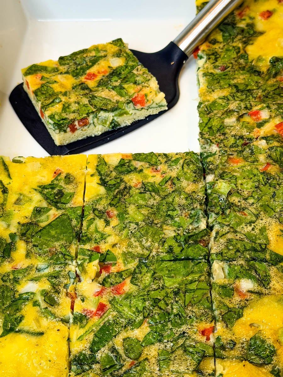 Baked spinach and vegetable eggs cut into squares in a baking dish, with one square lifted out on a spatula.