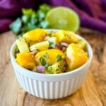 Fresh pineapple mango salsa with red onion and cilantro served in a white bowl with lime and cilantro in the background.