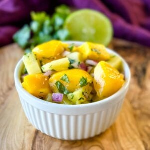 Fresh pineapple mango salsa with red onion and cilantro served in a white bowl with lime and cilantro in the background.