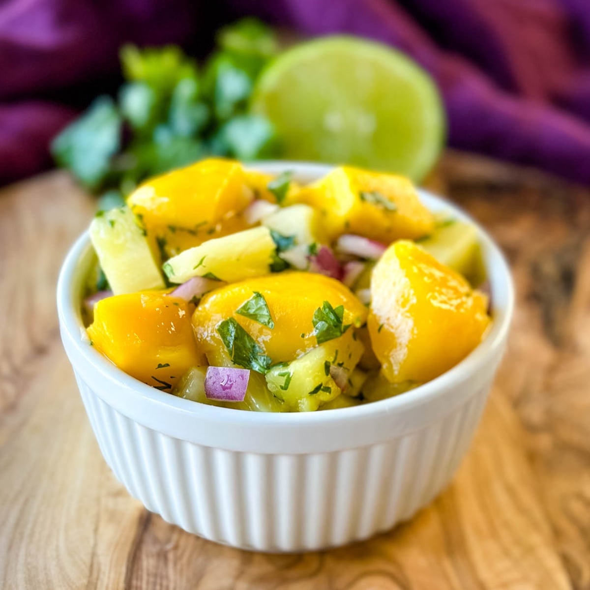 Fresh pineapple mango salsa with red onion and cilantro served in a white bowl with lime and cilantro in the background.