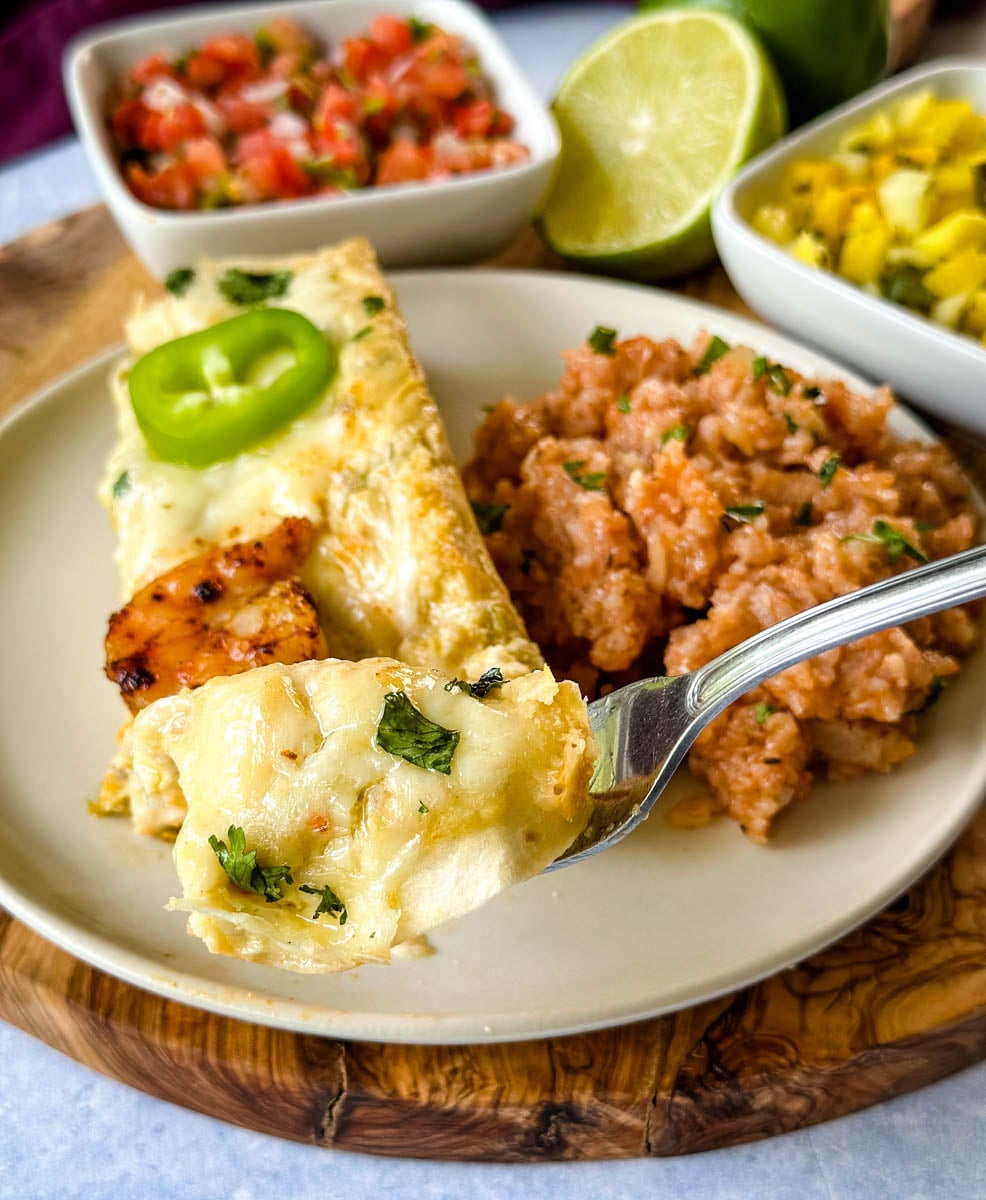 Creamy shrimp enchiladas served with Mexican rice, pico de gallo, and a fresh salsa, with a fork holding a cheesy bite of enchilada in the foreground.
