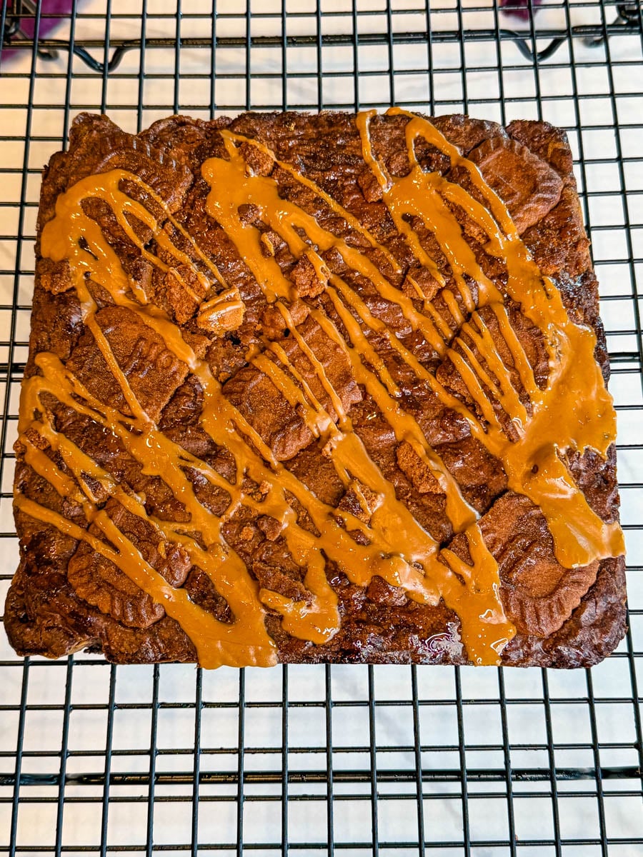 Freshly baked Biscoff brownies resting on a wire cooling rack, topped with Biscoff cookie pieces and a generous drizzle of melted cookie butter.