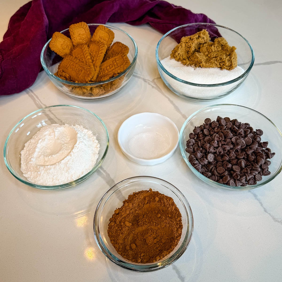 Biscoff brownie dry ingredients arranged in glass bowls, including Biscoff cookies, granulated and brown sugar, flour, salt, chocolate chips, and unsweetened cocoa powder.