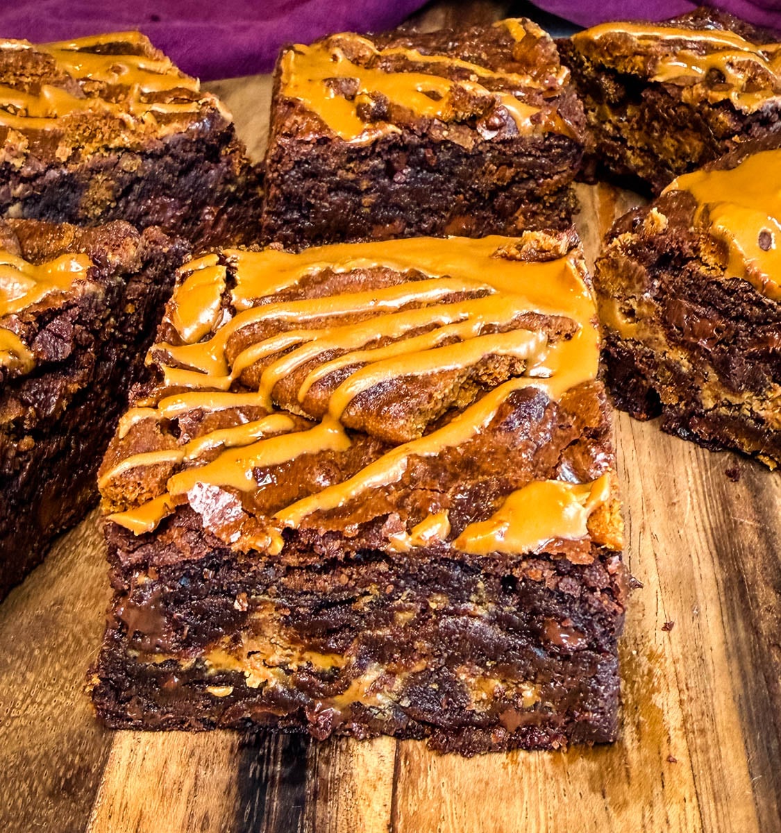 Close-up of thick, fudgy Biscoff brownies on a wooden board with visible chocolate chips, cookie butter swirls, Biscoff cookie pieces, and melted cookie butter drizzled over the top.