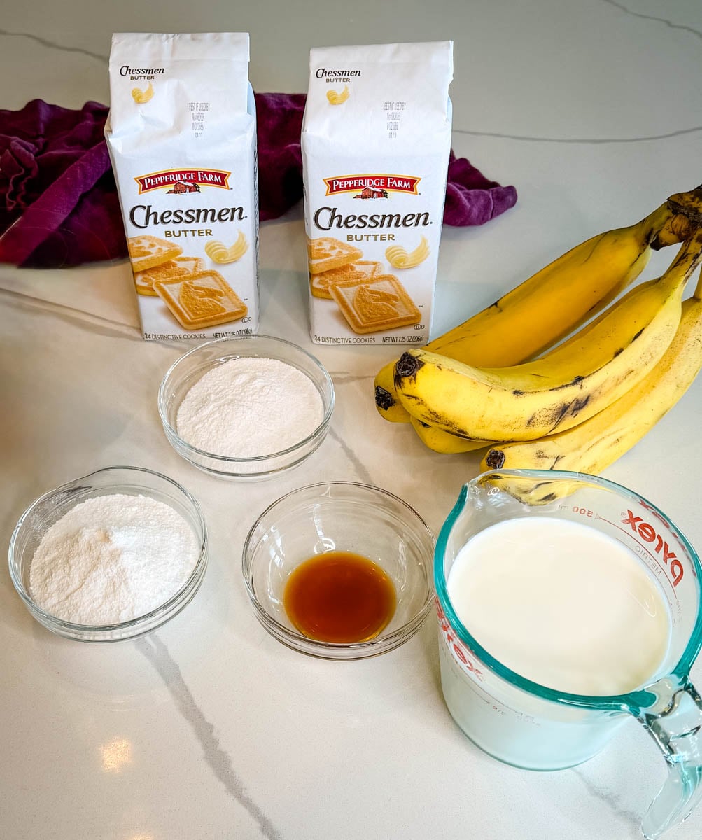 Ingredients for Chessman banana pudding, including Chessman butter cookies, ripe bananas, milk, vanilla, and pudding ingredients arranged on a countertop.