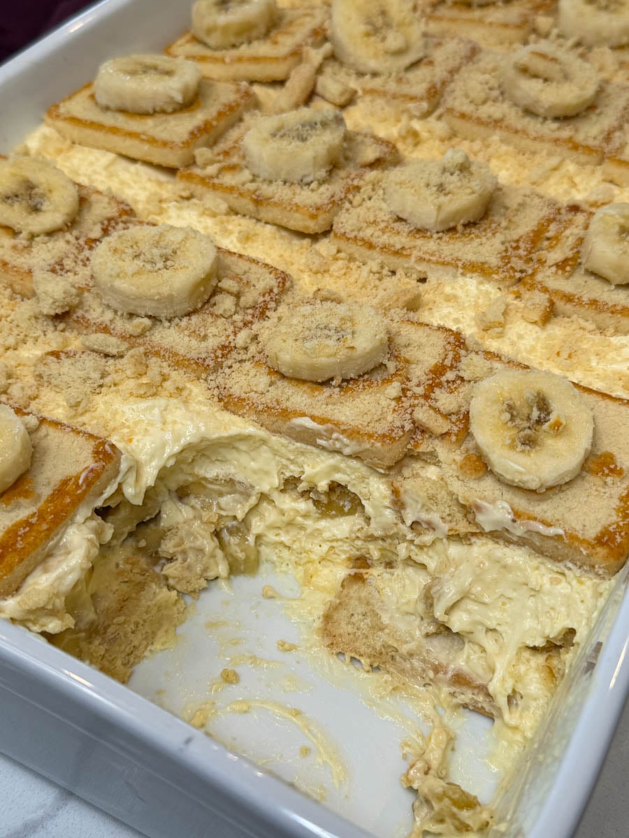 Close-up of a partially served pan of Chessman banana pudding showing creamy pudding, banana slices, cookie layers, and crushed cookies.