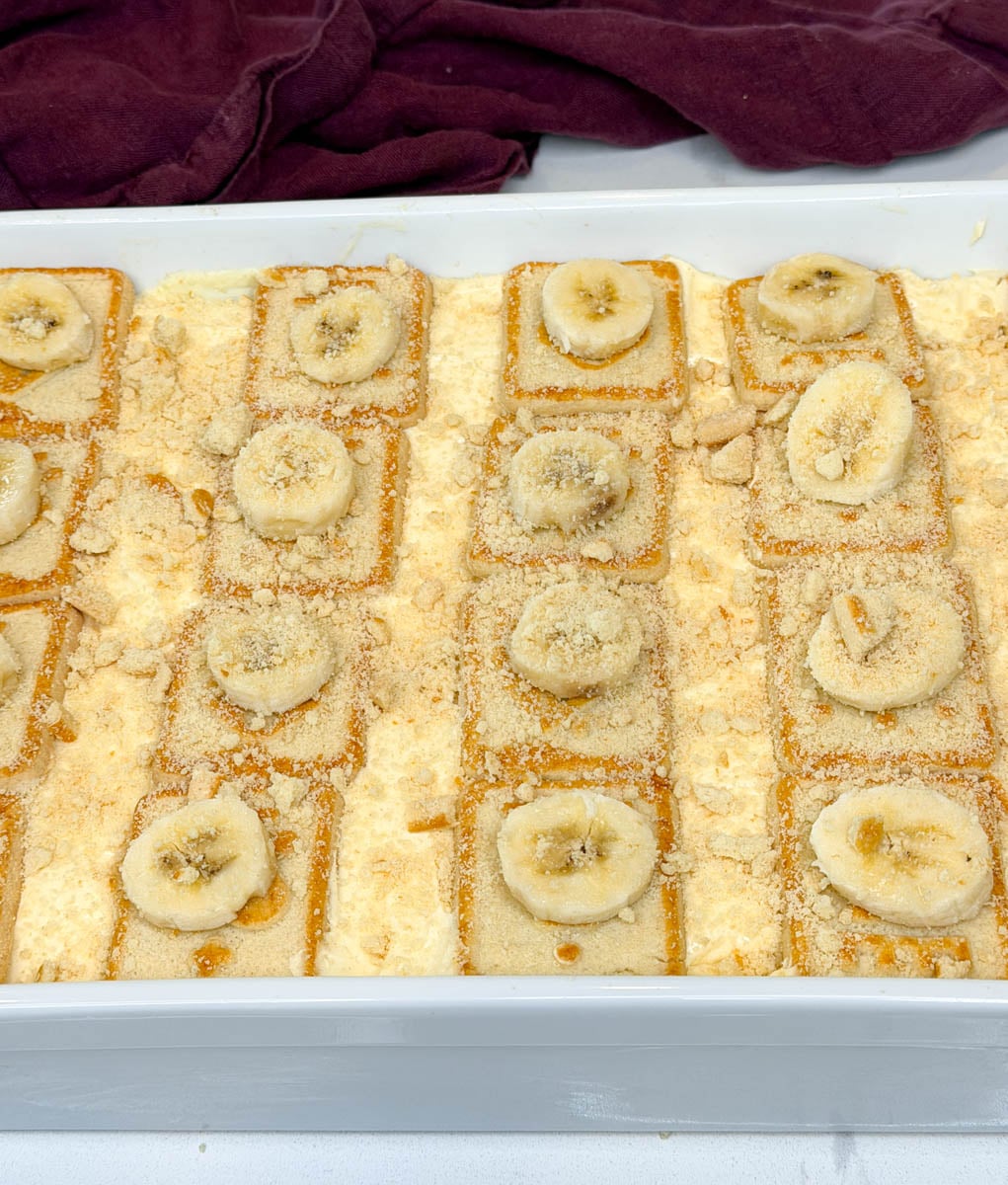 Pan of homemade Chessman banana pudding topped with rows of Chessman cookies, fresh banana slices, and crushed cookie crumbs.